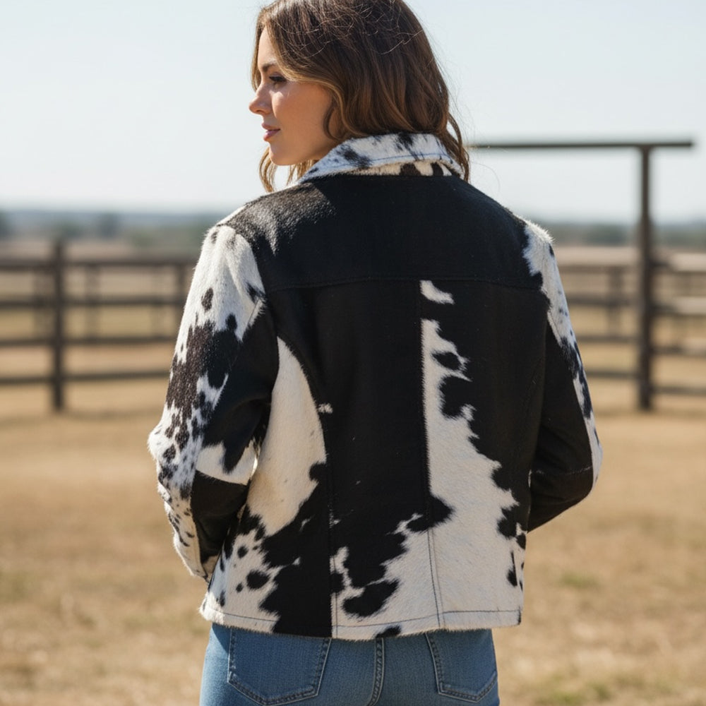 A woman wearing a black and white cow-print jacket, shown from the back in an outdoor ranch setting with a fence in the background.