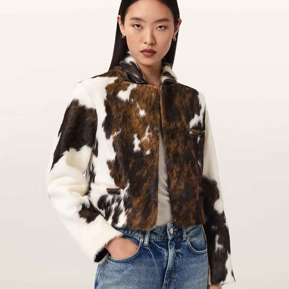 A model wearing a cropped cow-print faux fur jacket with brown, black, and white patches, styled with a white top and blue jeans against a plain light background.