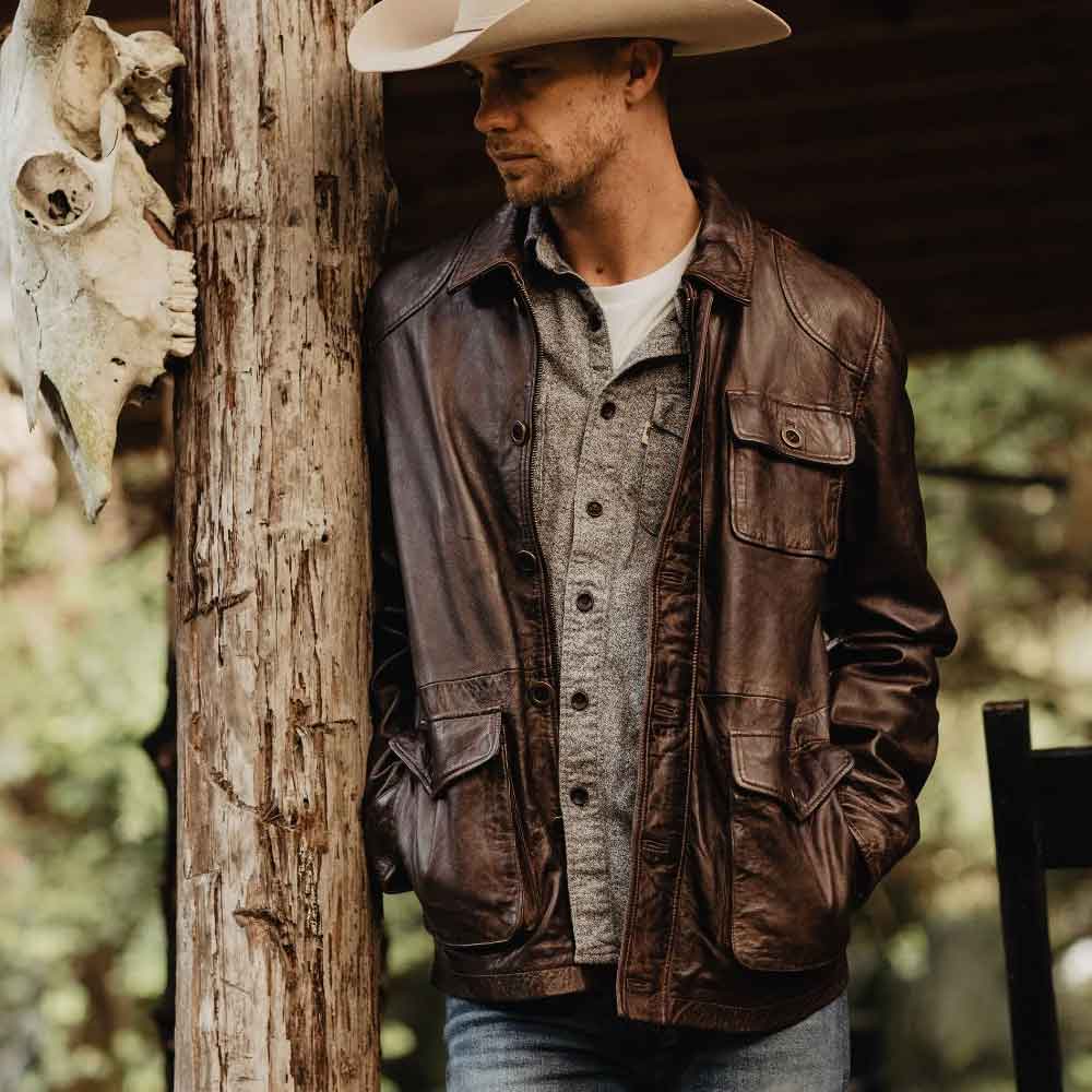 Man wearing a brown leather jacket and cowboy hat leaning against a tree with a blurred natural background