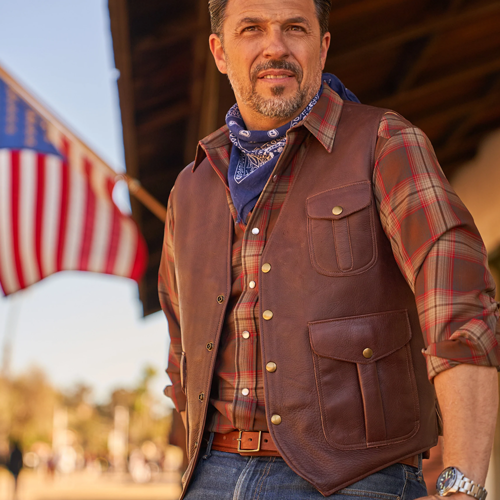 Man wearing a brown leather vest and plaid shirt with an American flag in the background