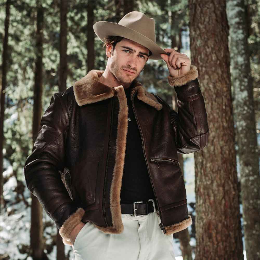 Man wearing a brown leather jacket with fur trim and a beige hat in a forest setting.