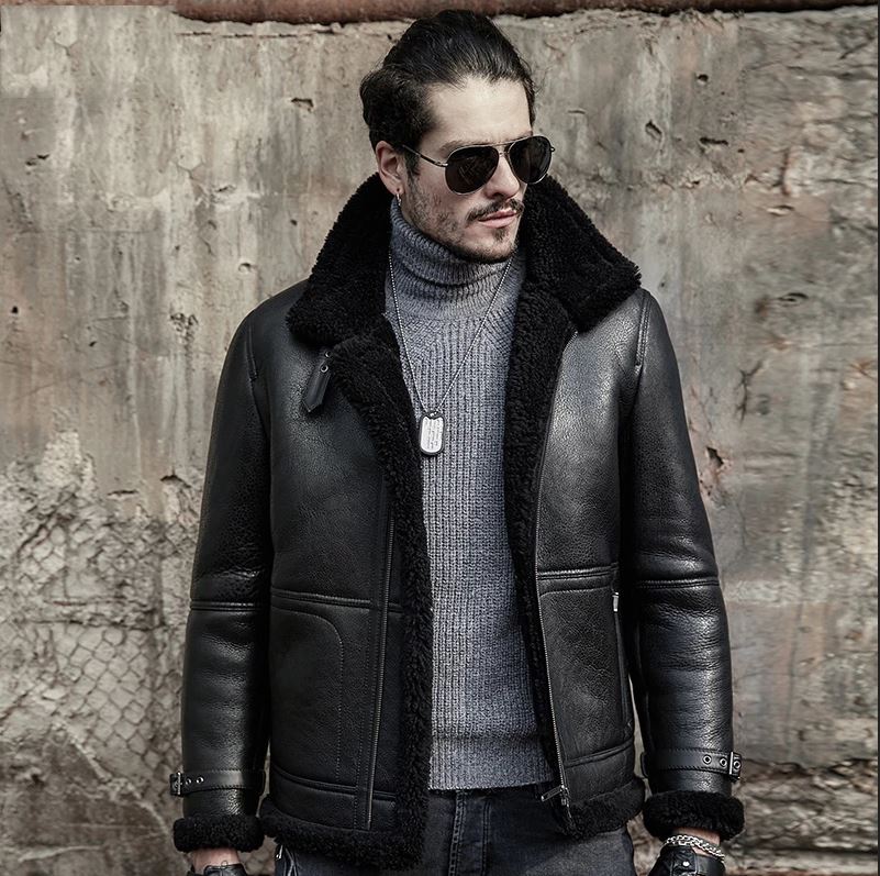 Man wearing a black leather jacket with fur collar and sunglasses against a textured wall.