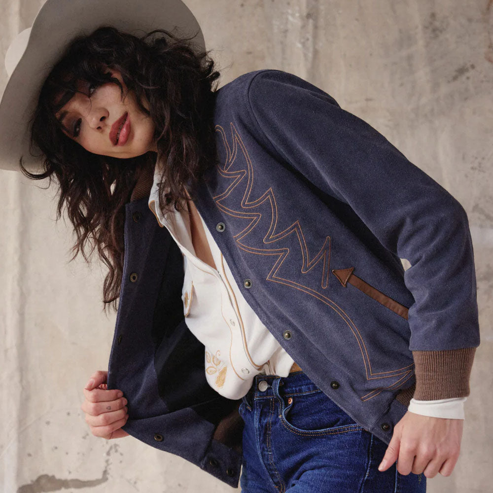 Person wearing a blue jacket with decorative stitching, white shirt, and jeans against a concrete wall.