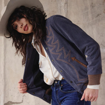 Person wearing a blue jacket with decorative stitching, white shirt, and jeans against a concrete wall.