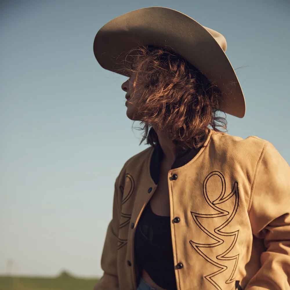 Person wearing a cowboy hat and tan jacket with a logo, standing against a clear sky.