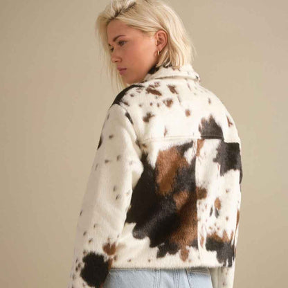 A woman wearing a fuzzy cropped jacket with a cow-print pattern in brown, black, and white, paired with light blue jeans, photographed against a plain beige background.