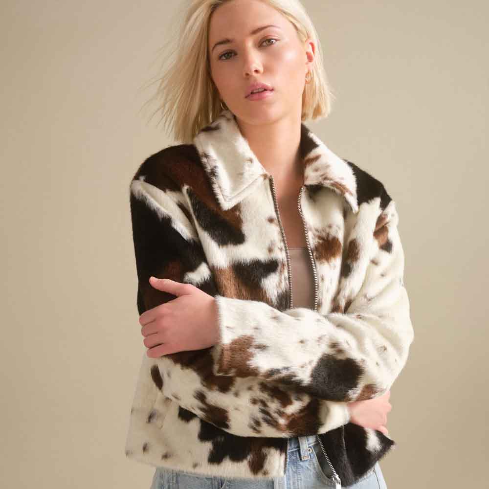 A woman wearing a brown and white cow-print faux fur jacket, standing against a plain beige background.