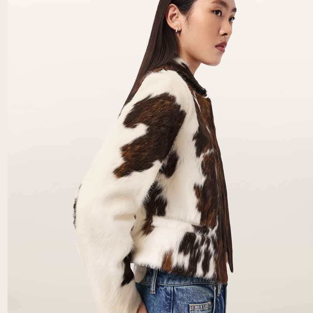 A woman shown in side profile wearing a cropped faux-fur jacket with a brown and white cow-print pattern, paired with blue jeans, against a plain light background.