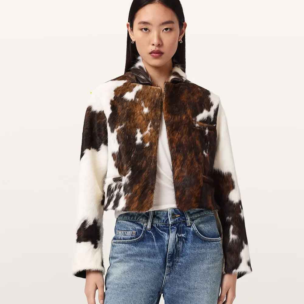 A model wearing a cropped faux-fur jacket with a brown and white cow-print pattern, styled with a white t-shirt and blue jeans against a plain off-white background.