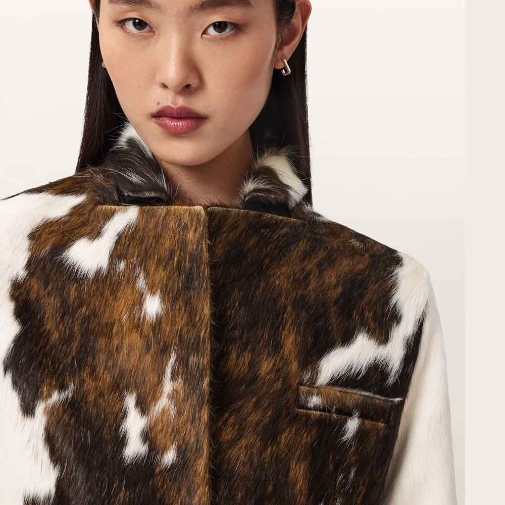 Close-up of a model wearing a brown and white cowhide faux fur vest with a matching faux fur collar, set against a plain light background.