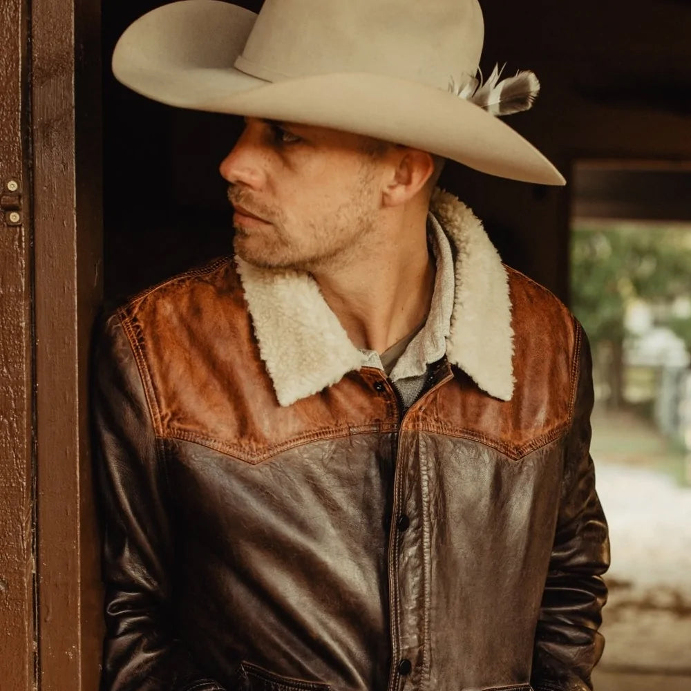 Man wearing a cowboy hat and brown leather jacket with white fur lining.