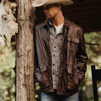 Man wearing a brown leather jacket and cowboy hat leaning against a tree with a blurred natural background