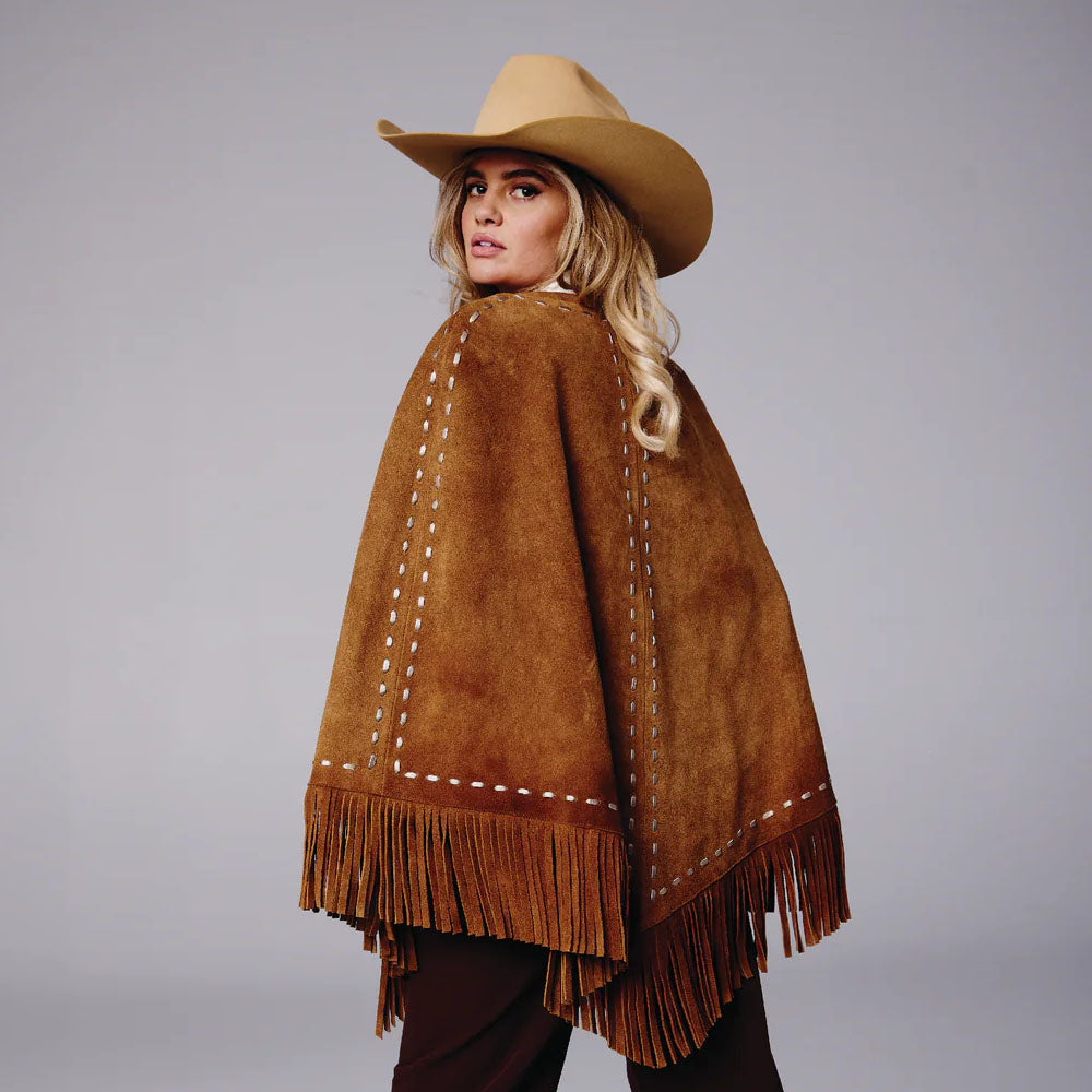 Woman wearing a brown suede poncho with fringe and a matching cowboy hat on a gray background