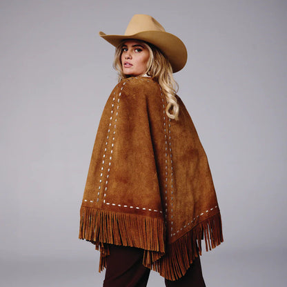 Woman wearing a brown suede poncho with fringe and a matching cowboy hat on a gray background