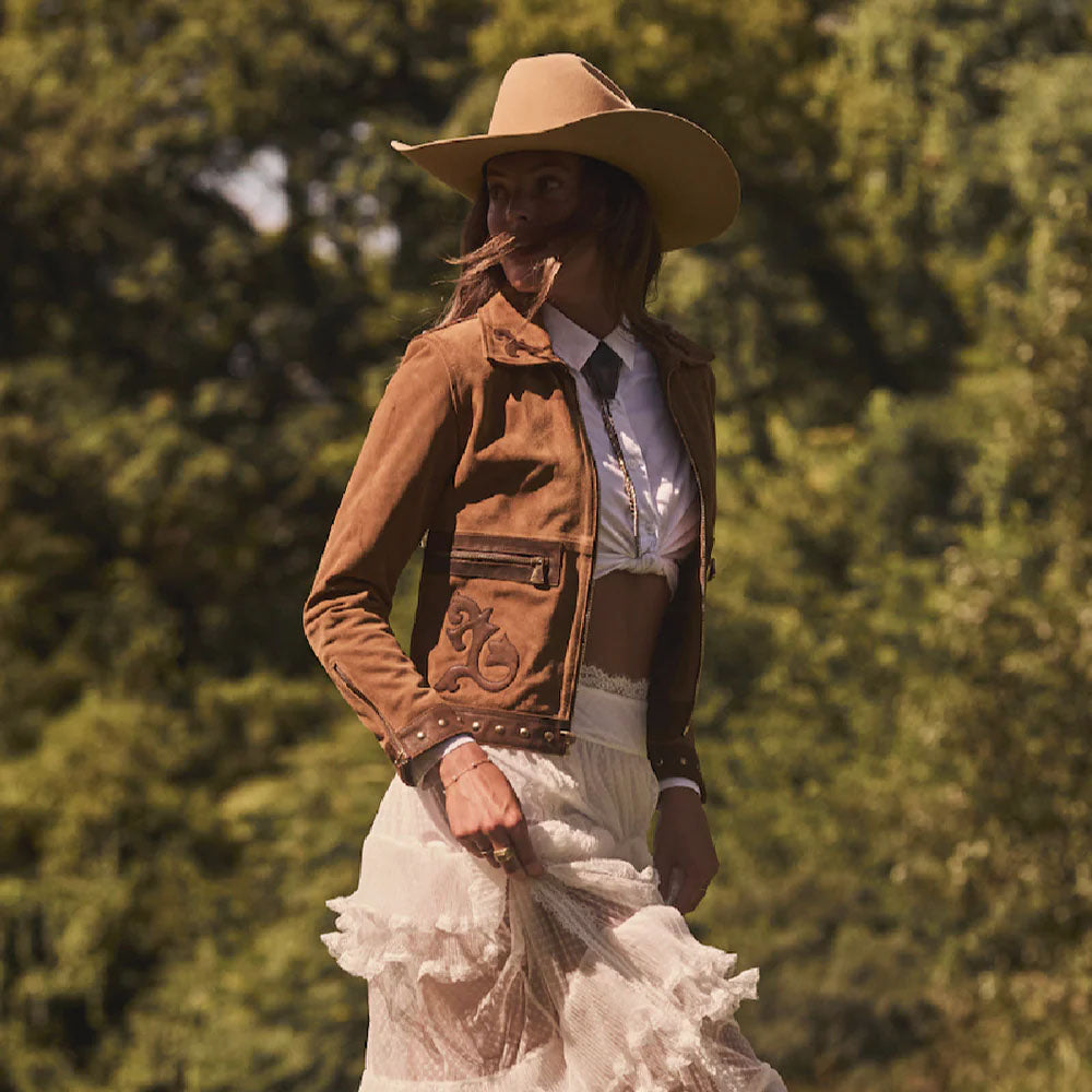 Person wearing a brown jacket, white dress, and cowboy hat with a blurred green background