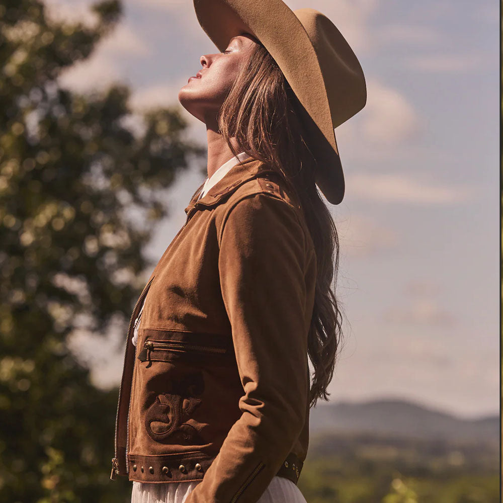 Woman wearing a brown hat and jacket against a scenic background with trees and mountains.