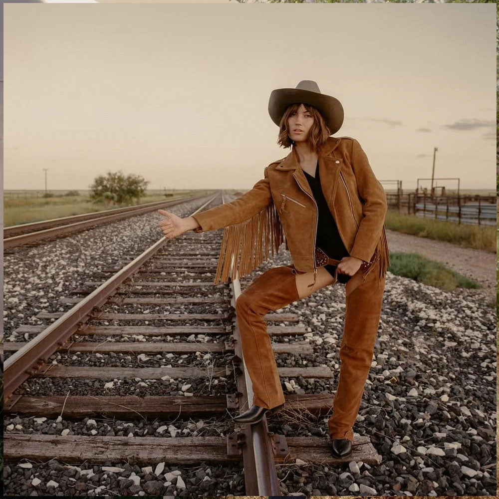 Person in a brown leather jacket and pants sitting on train tracks in a desert setting