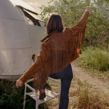 Person wearing a brown fringed jacket standing next to a vintage camper in a natural setting.