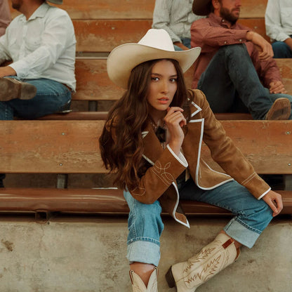 Woman in cowboy hat and boots sitting on bleachers with people in the background