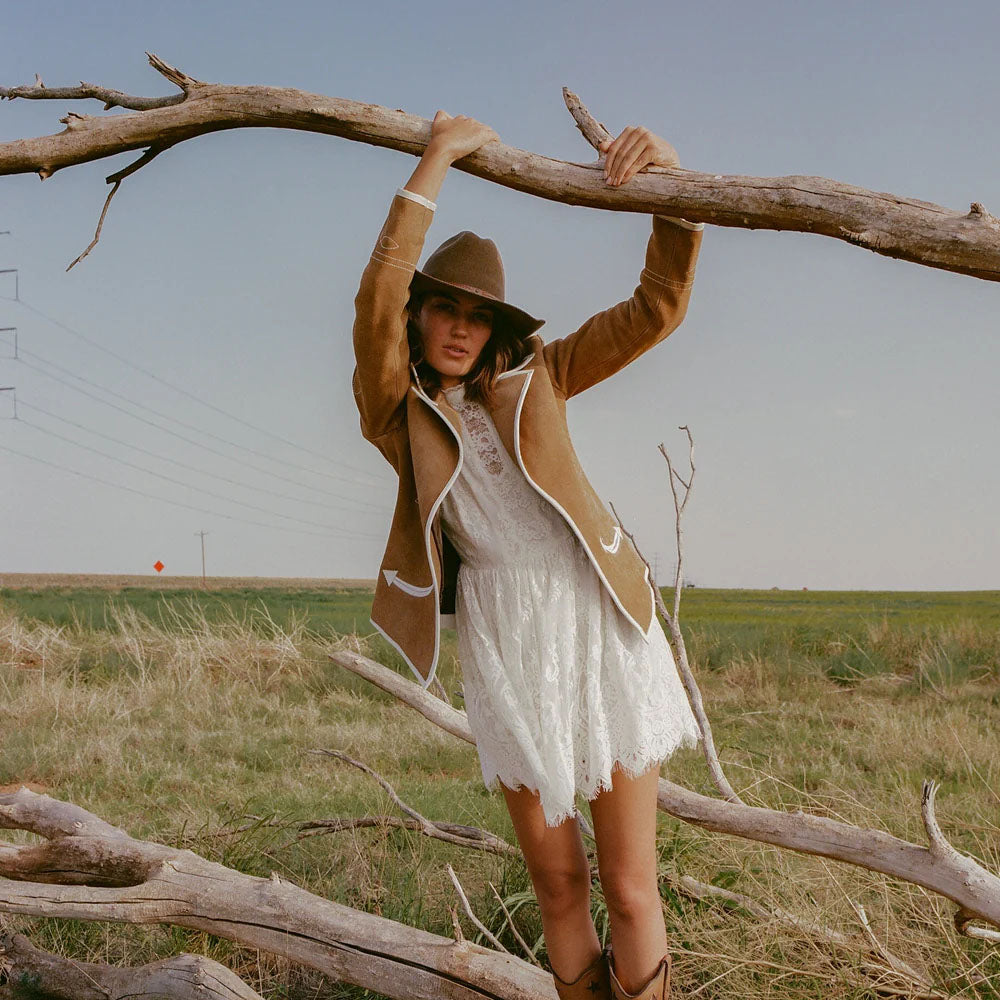 Woman in a white dress and brown jacket standing among fallen branches in an open field.