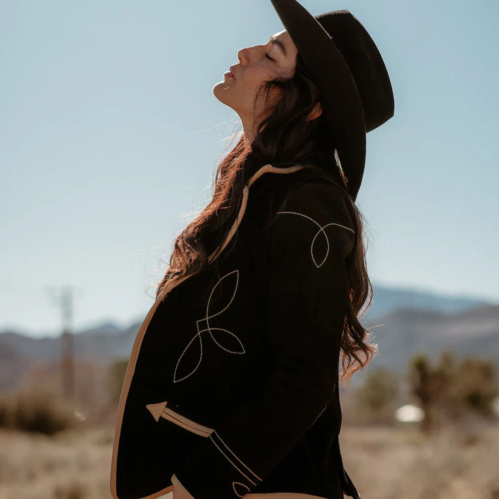 Woman wearing a black coat with white patterns and a hat, standing in a desert landscape.