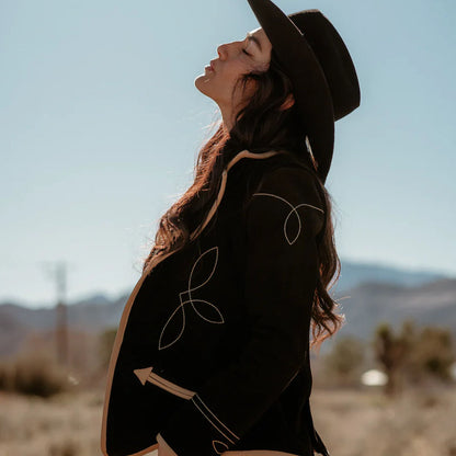 Woman wearing a black coat with white patterns and a hat, standing in a desert landscape.