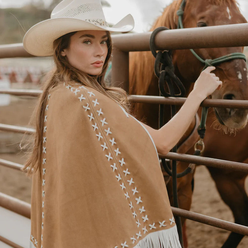 Woman in a cowboy hat and brown poncho standing next to a horse in a corral.
