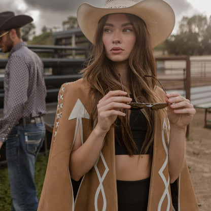 Woman in a cowboy hat and patterned coat holding a small object in a rural setting.