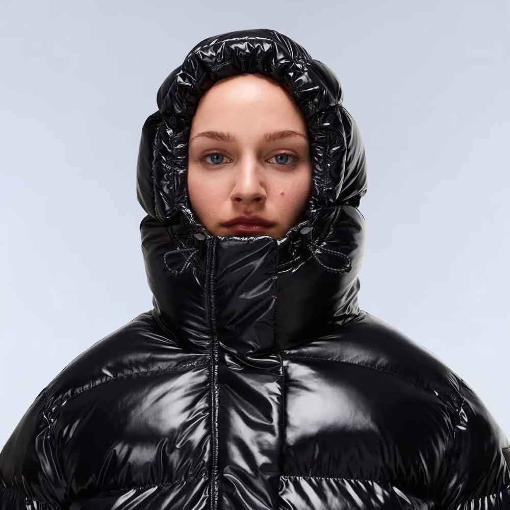 A close-up of a model wearing a shiny black puffer jacket with a high, gathered hood against a light gray studio background.