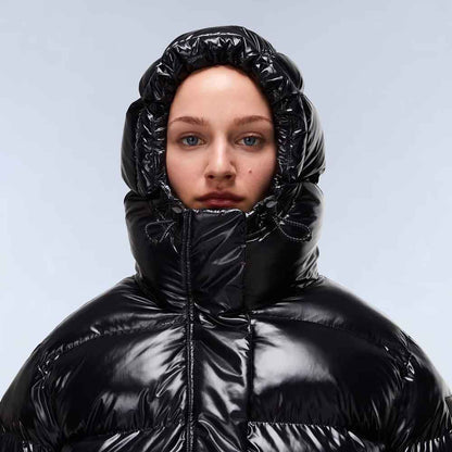 A close-up of a model wearing a shiny black puffer jacket with a high, gathered hood against a light gray studio background.