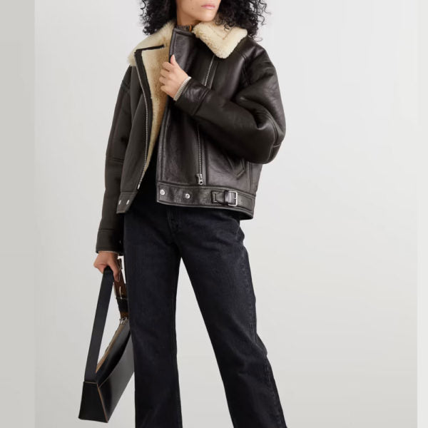Person wearing a dark leather jacket with shearling collar and holding a black handbag on a light gray background