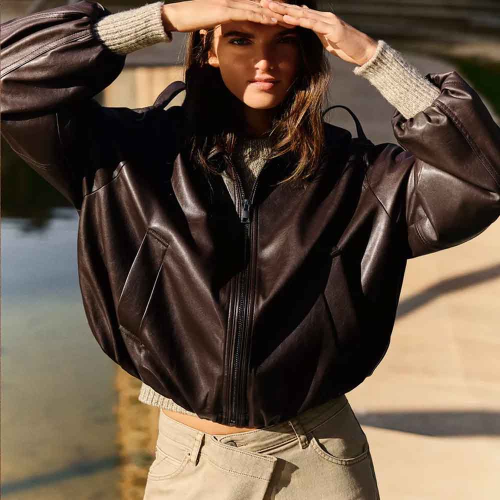A woman wearing a dark brown leather jacket with ribbed knit cuffs, posing outdoors with her hands raised to shield her eyes.
