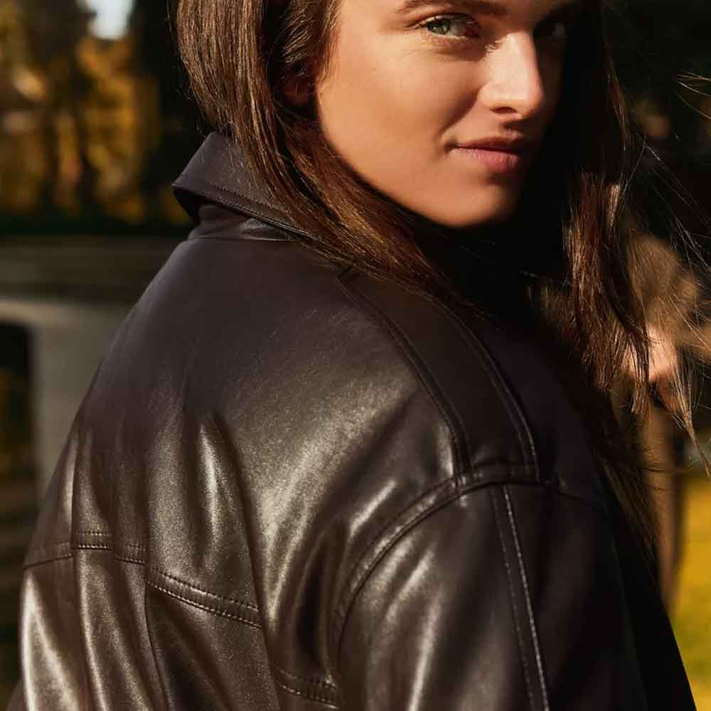 A close-up lifestyle shot of a woman wearing a dark brown leather jacket, looking back over her shoulder in warm sunlight.