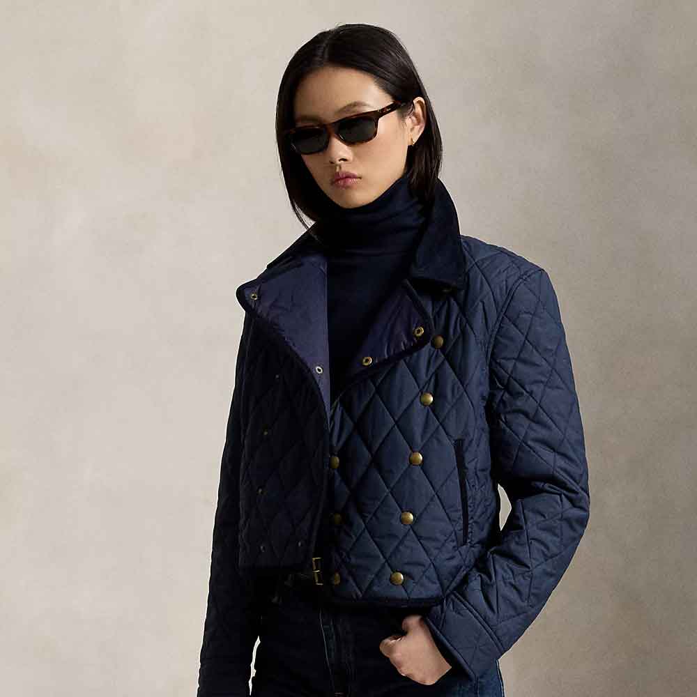 A model wearing a navy blue quilted jacket with gold-tone snap buttons over a dark navy turtleneck, styled with black sunglasses and dark jeans against a textured light gray background.