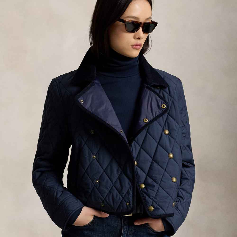 A woman wearing a navy blue quilted jacket with gold-tone snap buttons over a dark navy turtleneck, styled with black sunglasses and dark jeans against a neutral beige background.