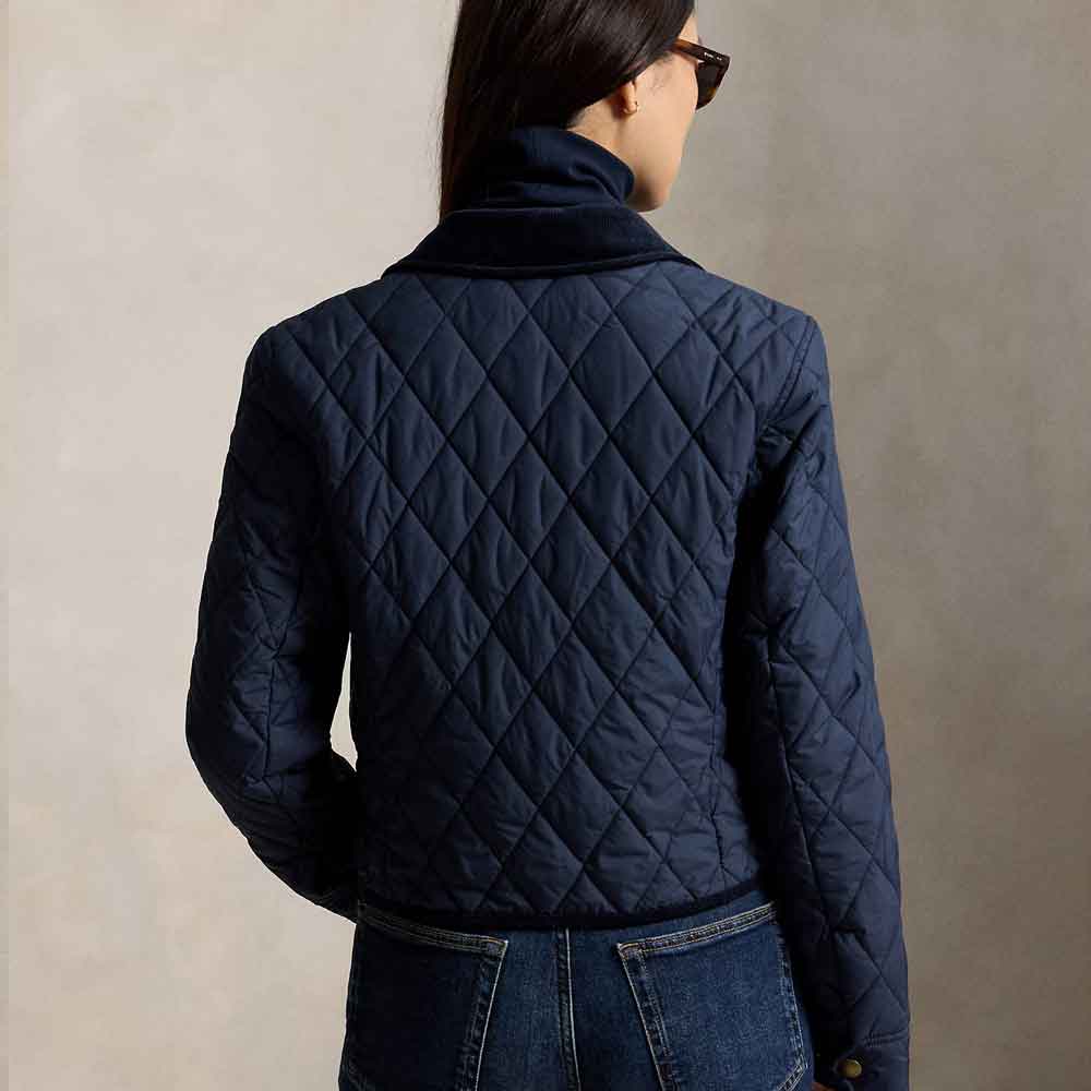 A woman shown from the back wearing a navy blue quilted jacket with a high collar, paired with blue jeans, against a neutral beige background.
