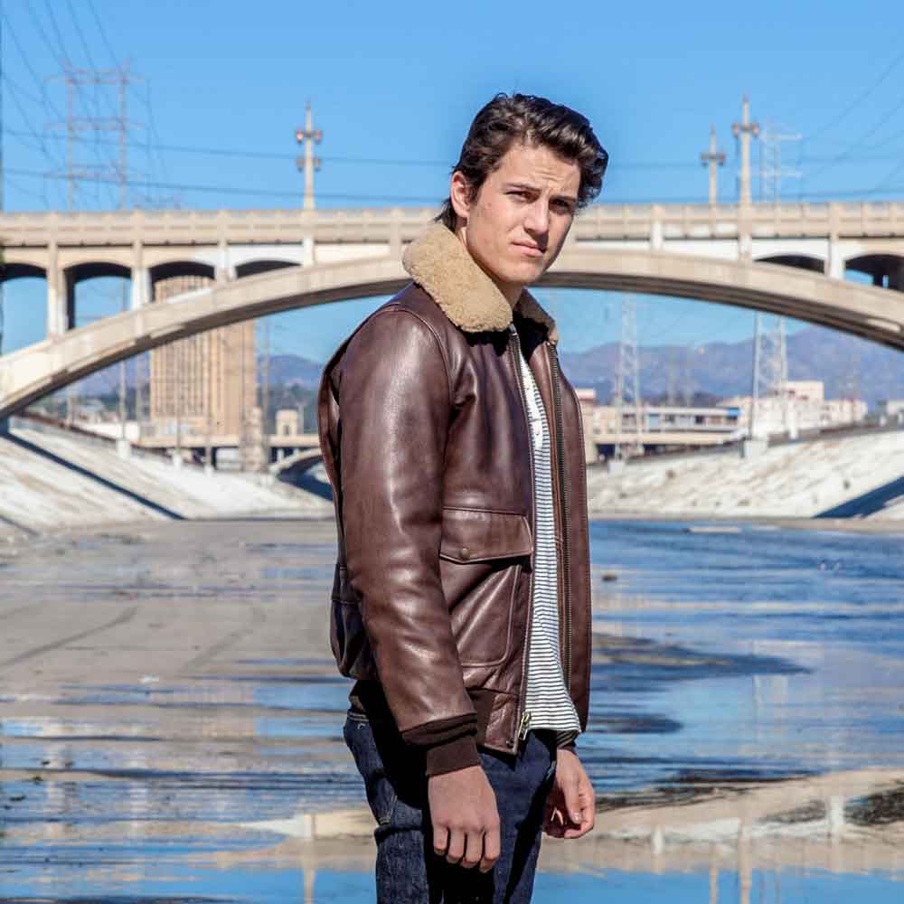 Man wearing a brown leather jacket with a fur-lined hood standing in front of a bridge.