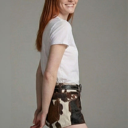 A woman wearing a plain white short-sleeve crewneck t-shirt and brown and white cow-print shorts with a black studded belt, shown from a side-back angle against a solid gray studio background.