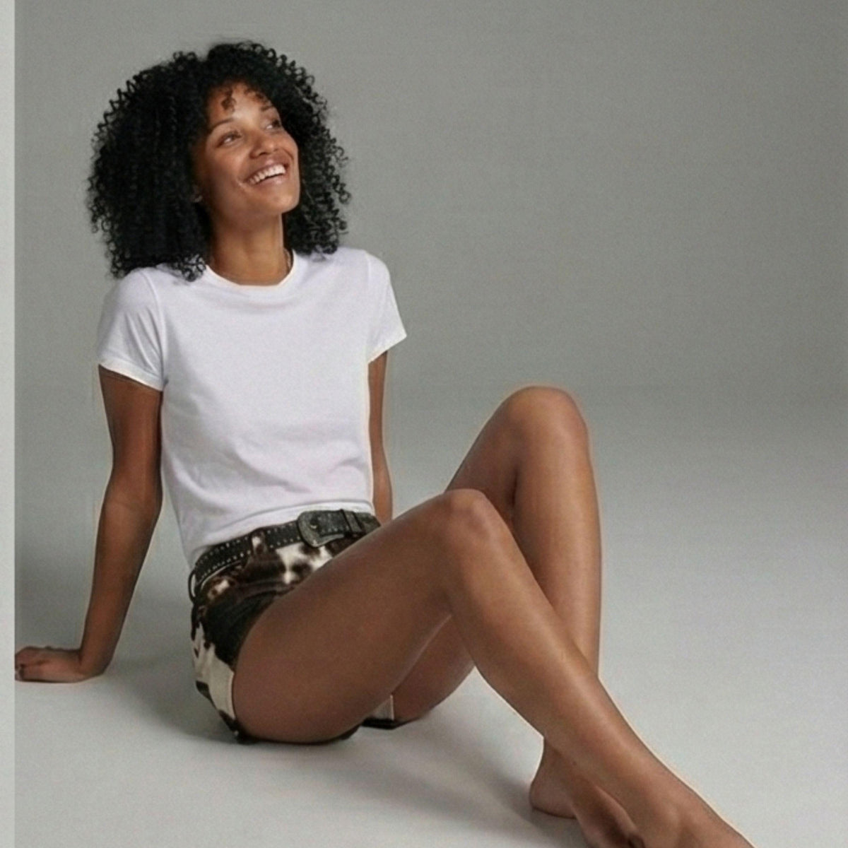 A woman with curly black hair sits on a light gray floor against a matching gray studio background, wearing a plain white short-sleeve T-shirt and dark patterned shorts.