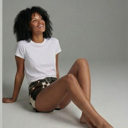 A woman with curly black hair sits on a light gray floor against a matching gray studio background, wearing a plain white short-sleeve T-shirt and dark patterned shorts.
