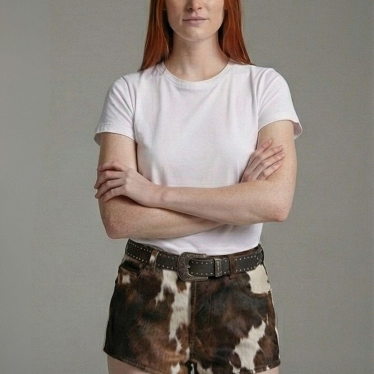A woman with red hair wearing a plain white short-sleeve crewneck t-shirt and brown and white cow-print shorts with a dark brown studded belt, standing with arms crossed against a solid gray studio background.