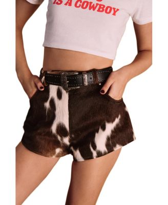 A person wearing brown and black cow-print shorts with a black belt, paired with a white cropped T-shirt that has red text reading 'IS A COWBOY'.