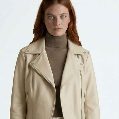 A woman with red hair wearing a light beige leather moto jacket over a taupe turtleneck, photographed against a clean white studio background.