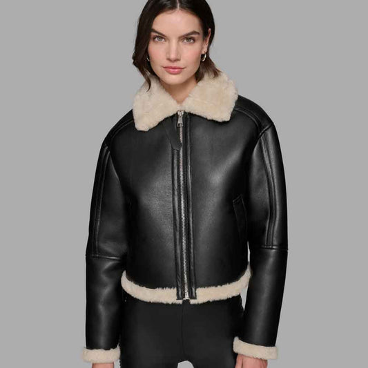 A woman wearing a black leather aviator jacket with a thick cream shearling collar, zipper front, and matching shearling trim at the hem and cuffs, photographed against a solid gray studio background.