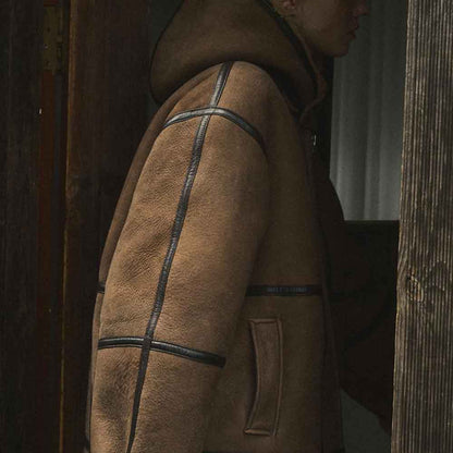 A person wearing a brown shearling-style hooded jacket with dark contrast stitching, shown in a dim wooden doorway setting.