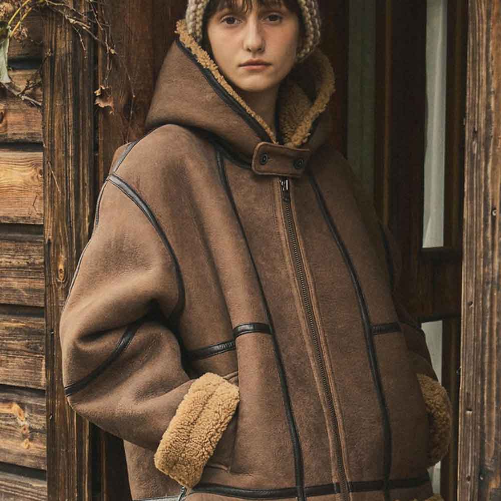 A person wearing a brown faux shearling-lined hooded jacket with black contrast stitching and a knit beanie, standing in front of rustic wooden doors.