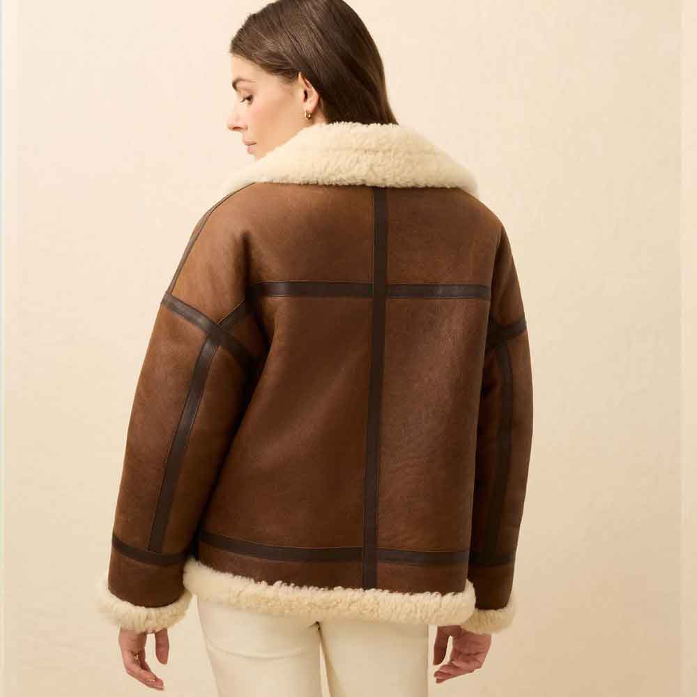 A woman shown from the back wearing a brown shearling jacket with dark contrast panels and a cream-colored collar and hem, paired with off-white pants, against a plain beige background.