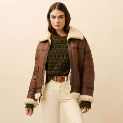A woman models a brown leather aviator jacket with a cream faux shearling collar and cuffs, worn over an olive green knit sweater and cream pants against a warm beige background.