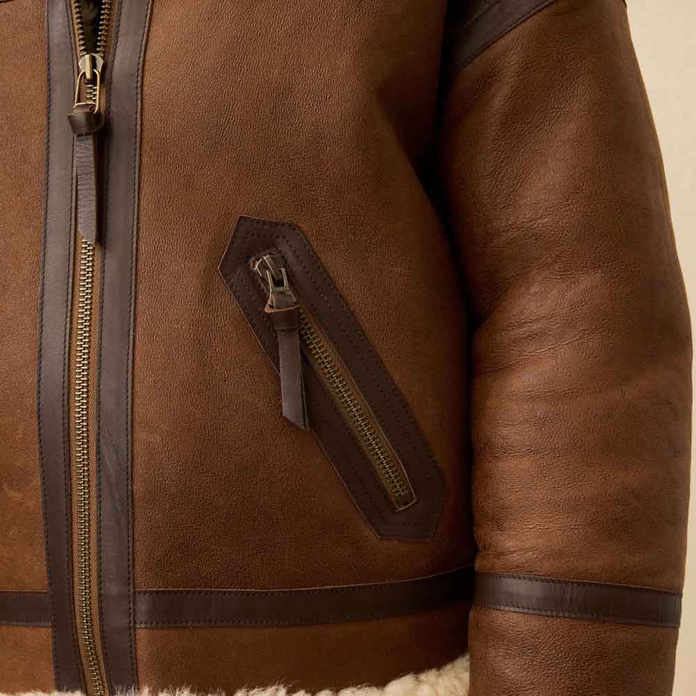 Close-up of a brown leather jacket showing the front zipper, a diagonal zippered pocket, and dark brown leather trim on a light beige background.