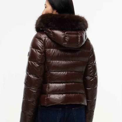 A woman viewed from the back wearing a glossy dark brown puffer jacket with a large dark fur-trimmed hood, paired with dark blue jeans, against a plain white background.